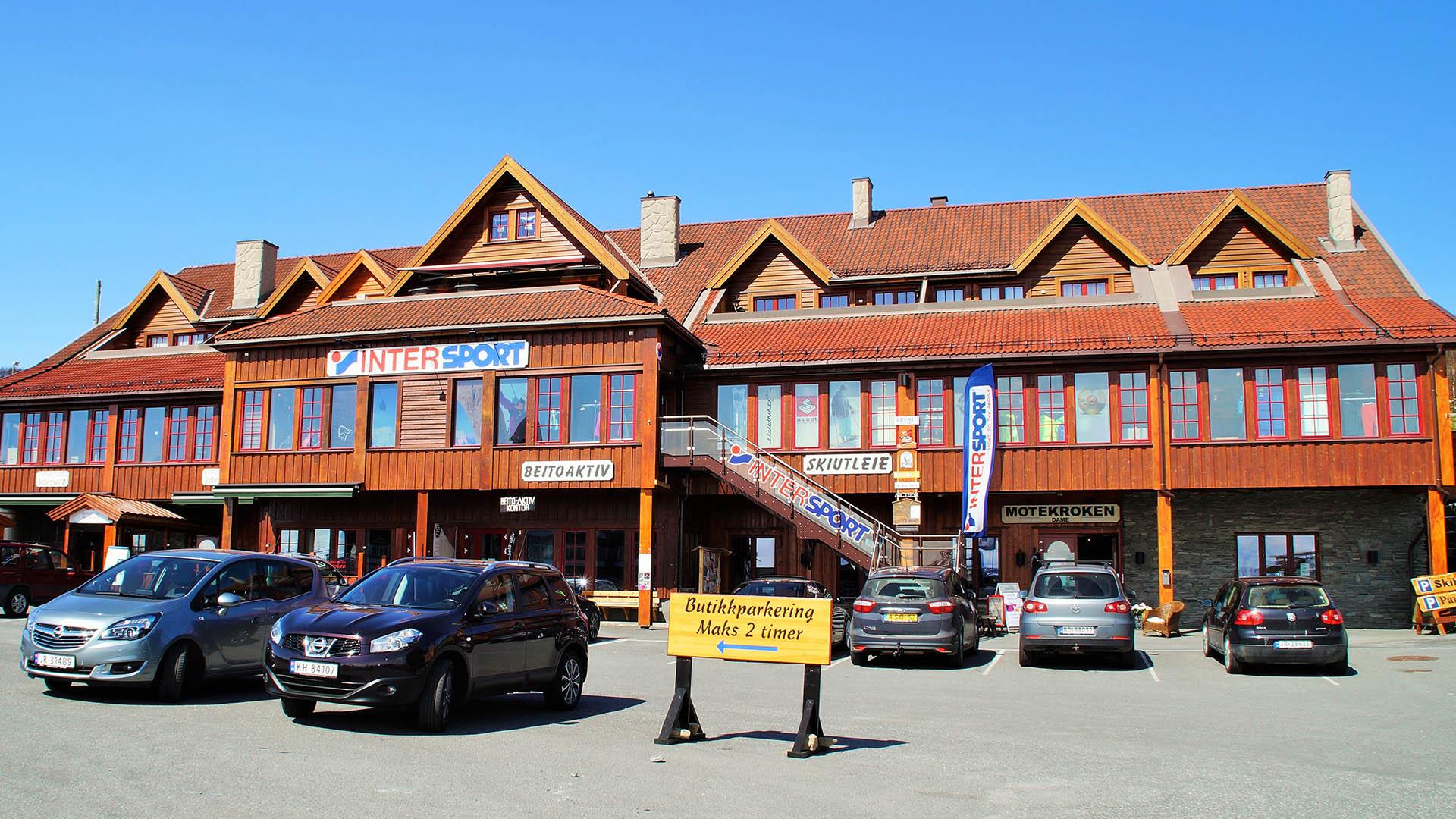 Light brown wooden building over three stories with several shops on a sunny day. Cars parked in front of it.