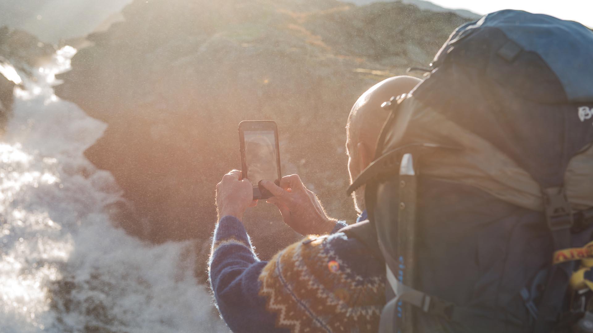 A mountain hiker with a large backpack holds up a smartphone to take a picture of a waterfall against the sun.