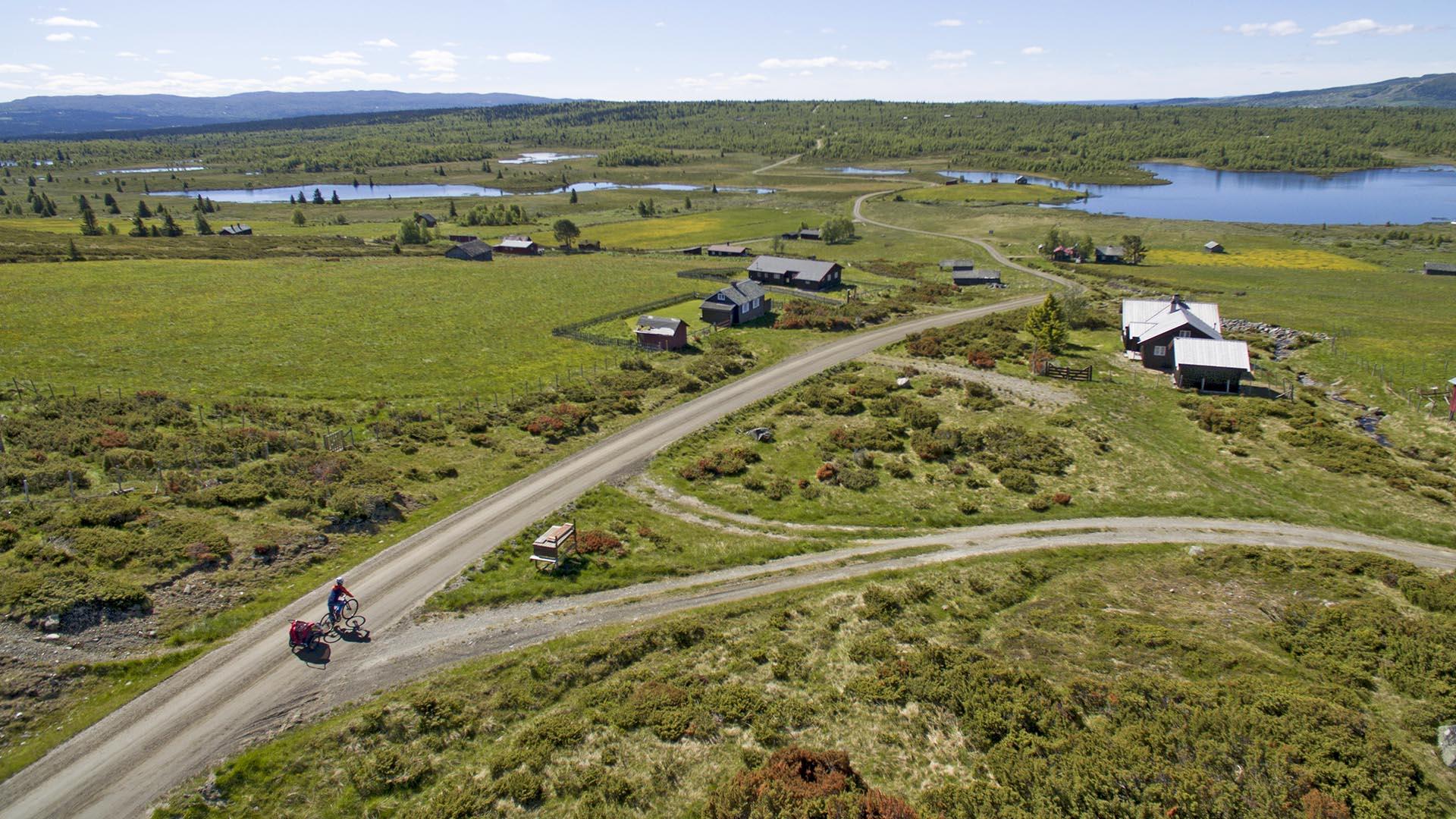 Drone image of a cyclist with trailer on a gravel road over an open high plateau with green pastures and some bushes. In the background a couple of small lakes.