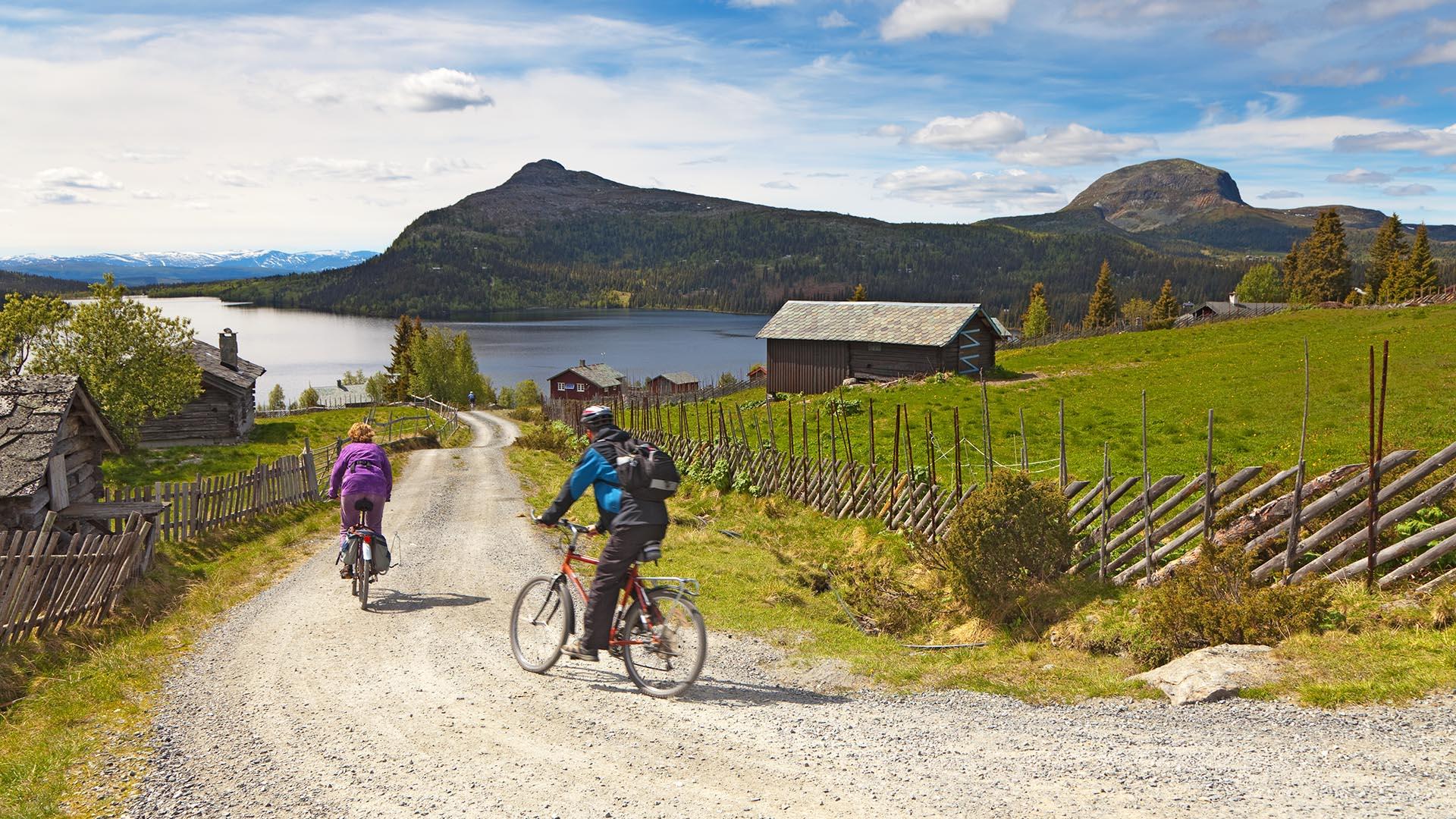 To syklister runder en sving på en stølsvei nedover mot et vann i idylliske omgivelser med skigard, en hytte på grønne stølsenger og fjell bakenfor vannet.