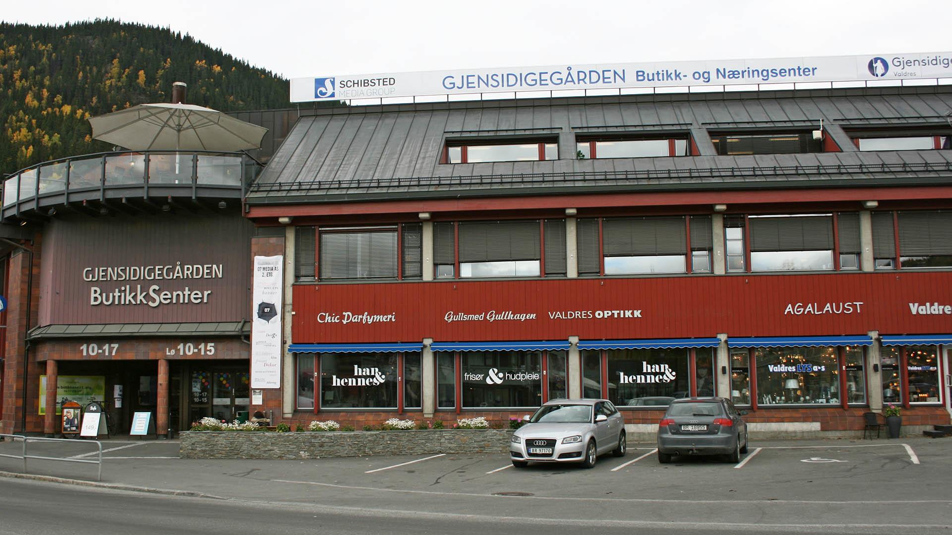 Image of a small mall with different shops and services. Cars are parked outside.