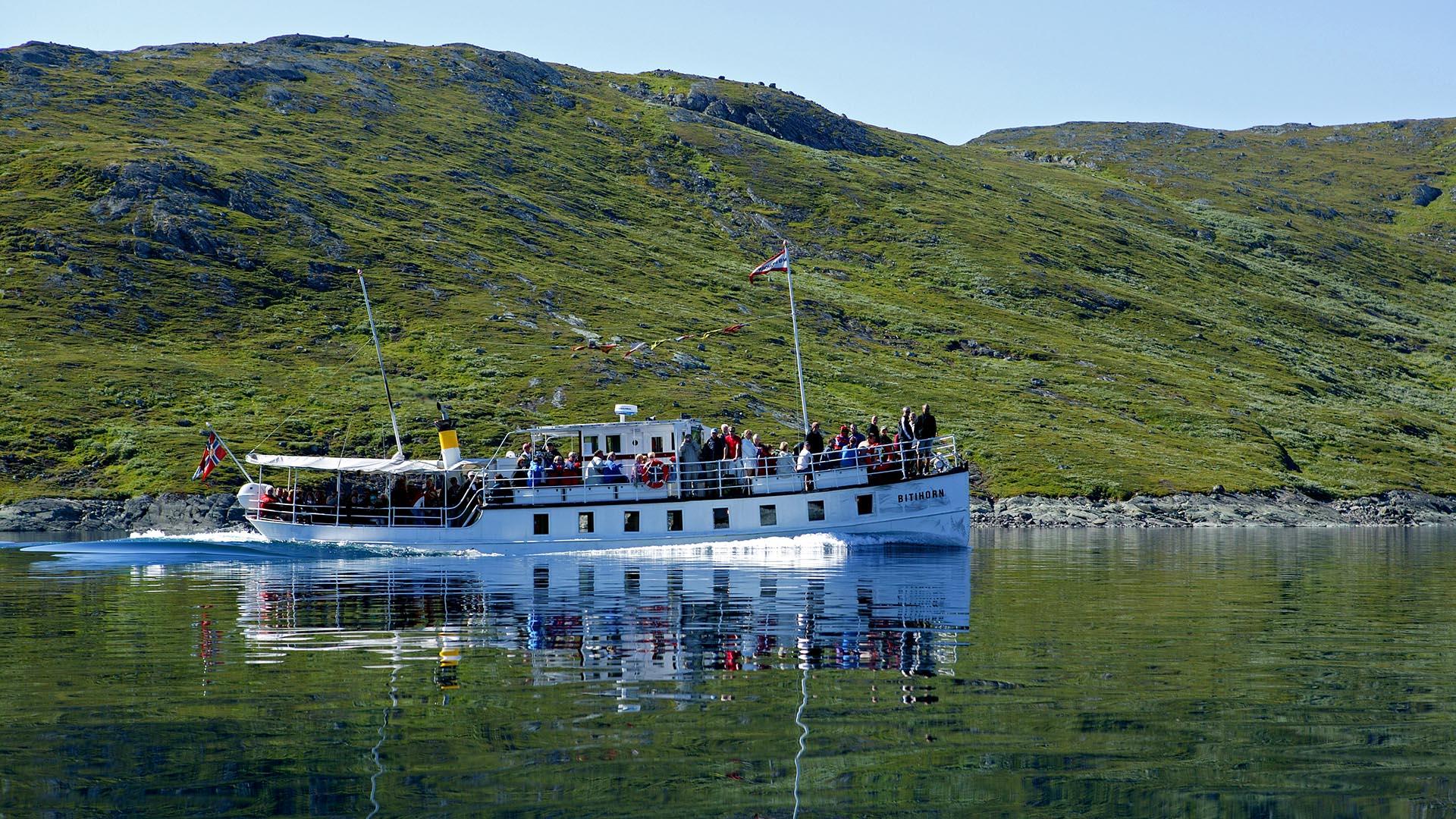 Hvit passasjerbåt på en fjellsjø. Passasjerer står på dekk. Fjell i bakgrunnen.