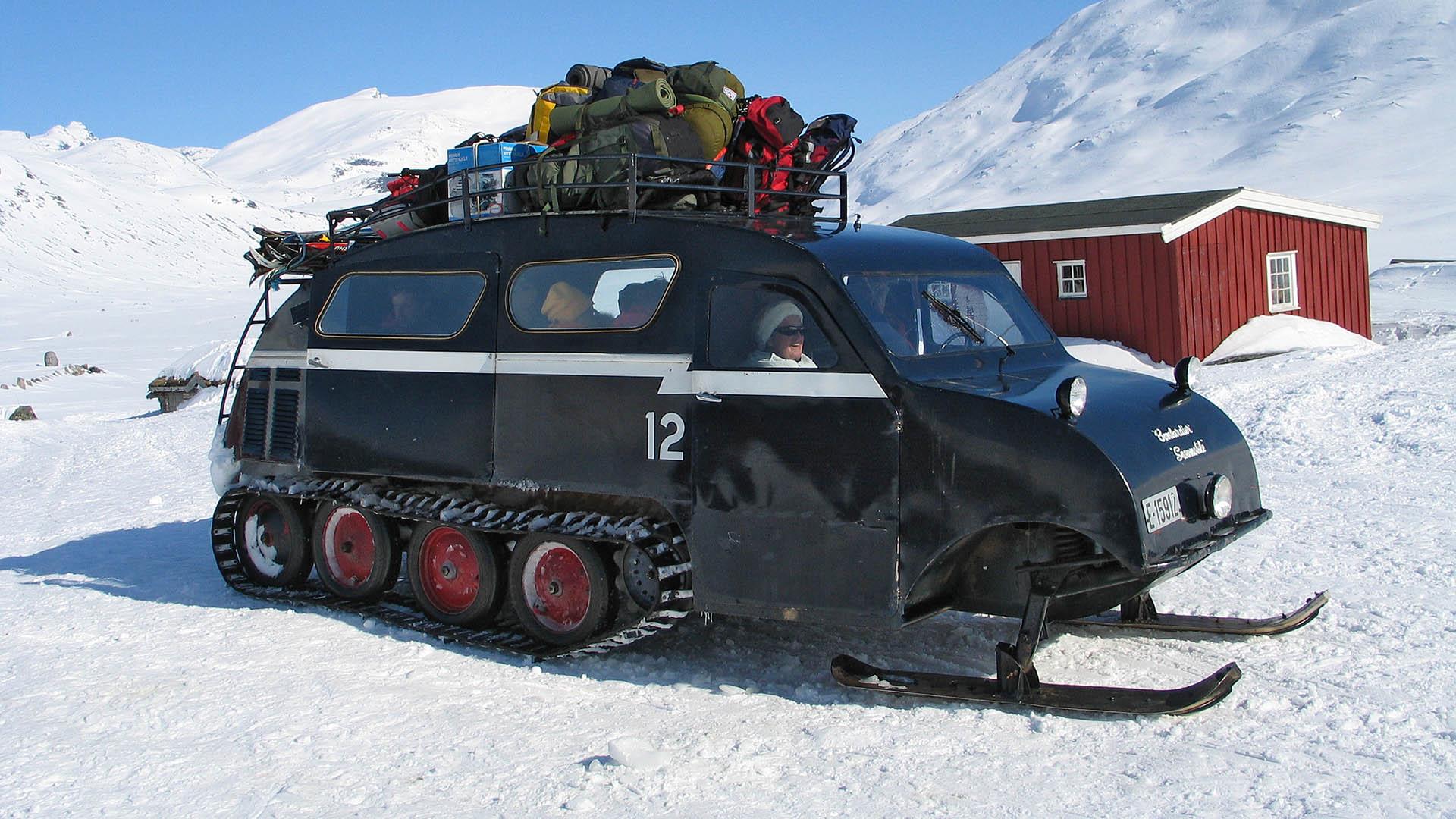 Beltebil med bagasje på taket parkert på snøen utenfor liten rød hytte.