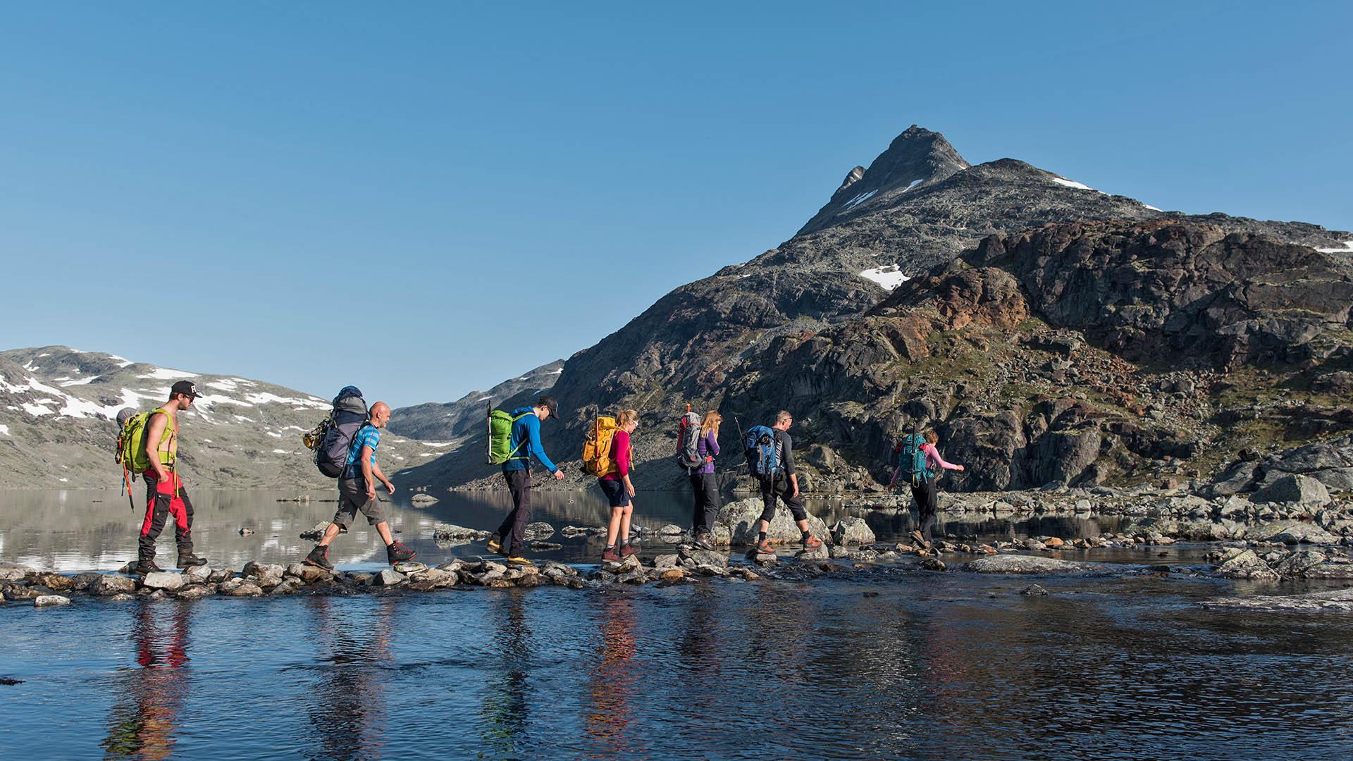 En gruppe fjellvandrere steingår en elv foran et fjell i goldt høyfjellslandskap.