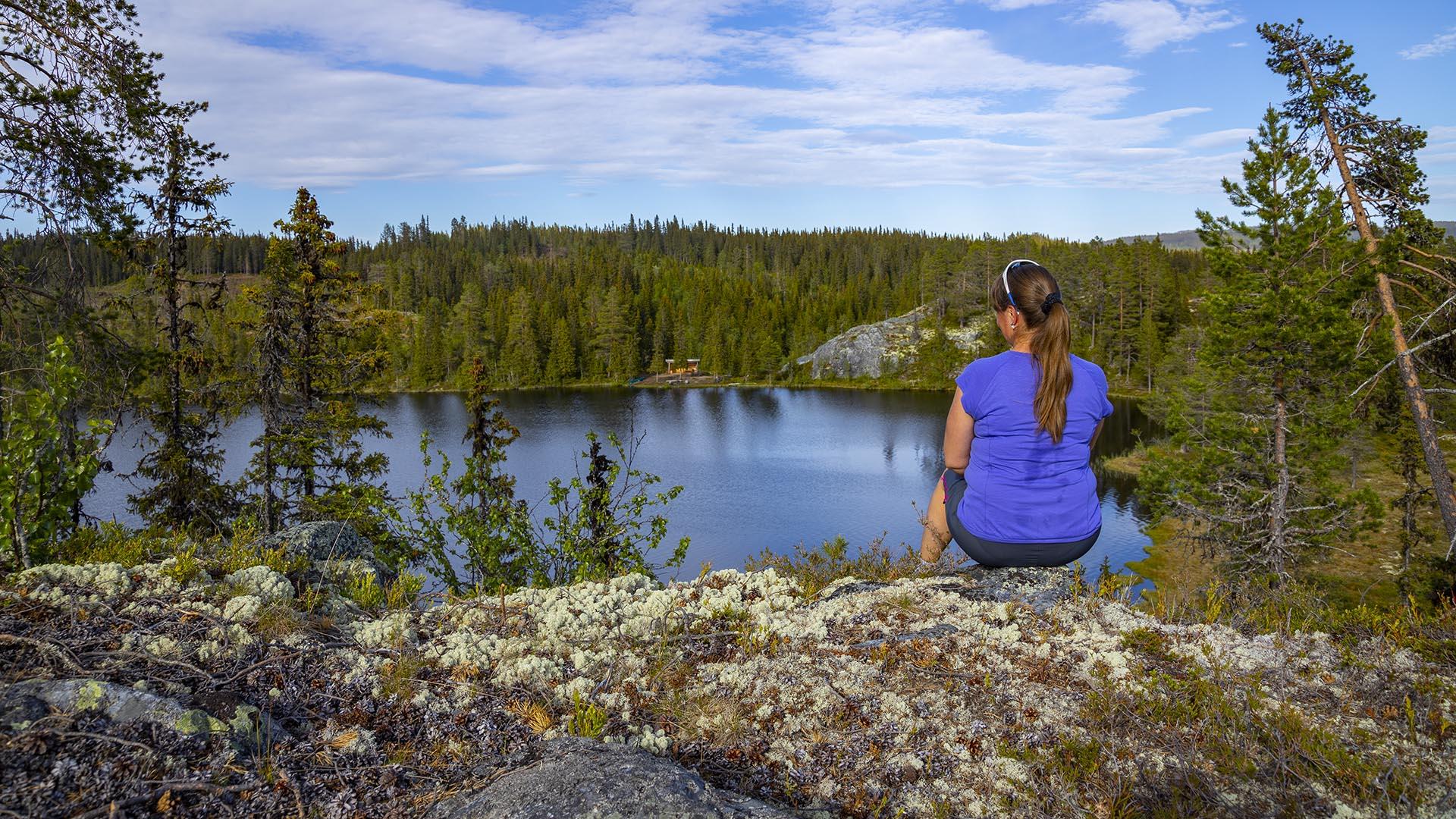 En dame i t-skjorte sitter på en lavbevokst fjellknaus ovenfor et skogsvann.