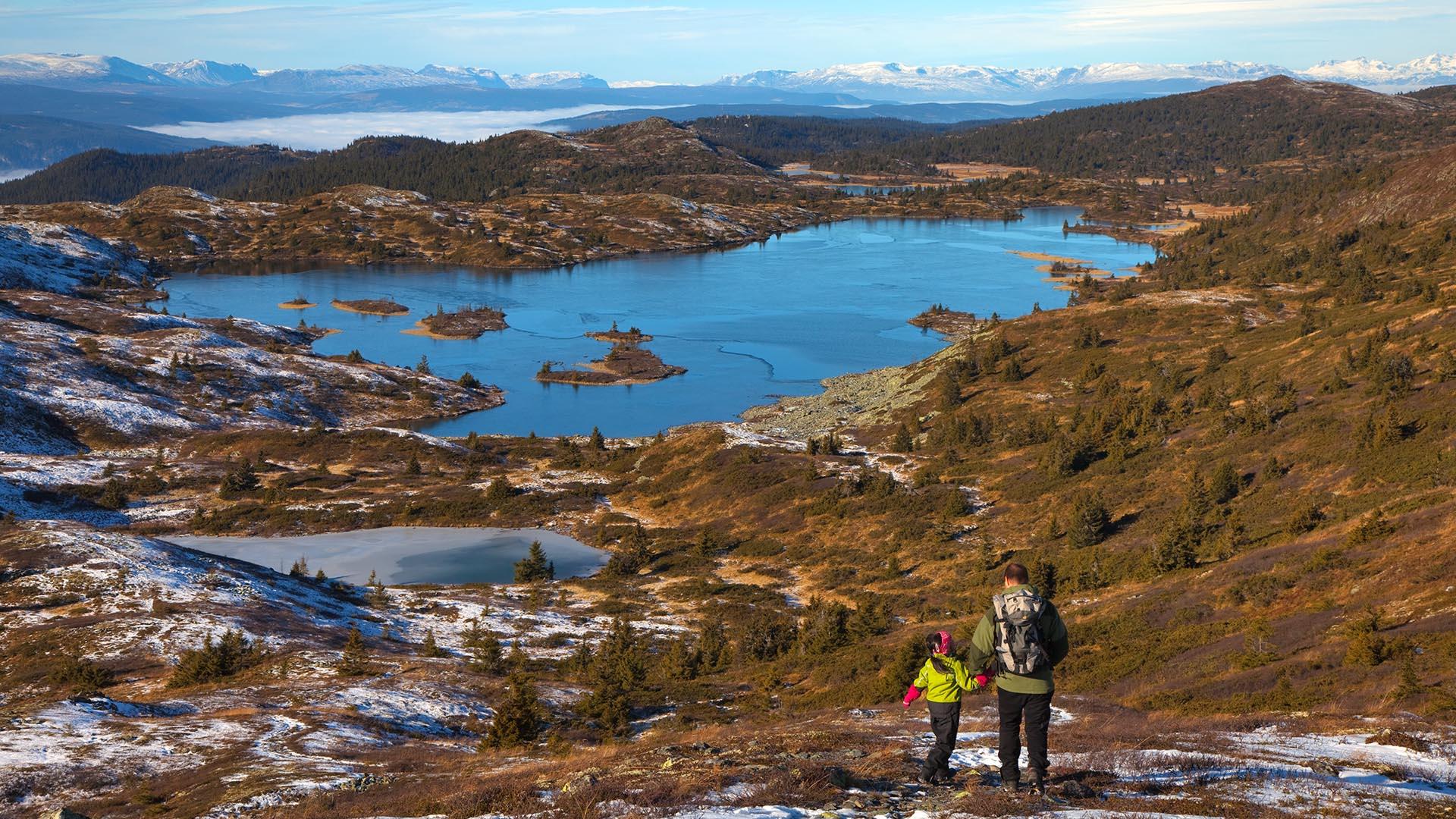 A man and a small girl in front of a great mountain landscape above the tree line with lakes and open, undulating country and high mountains on the far horizon. Patches of the first snow have endured the sun in shady places.