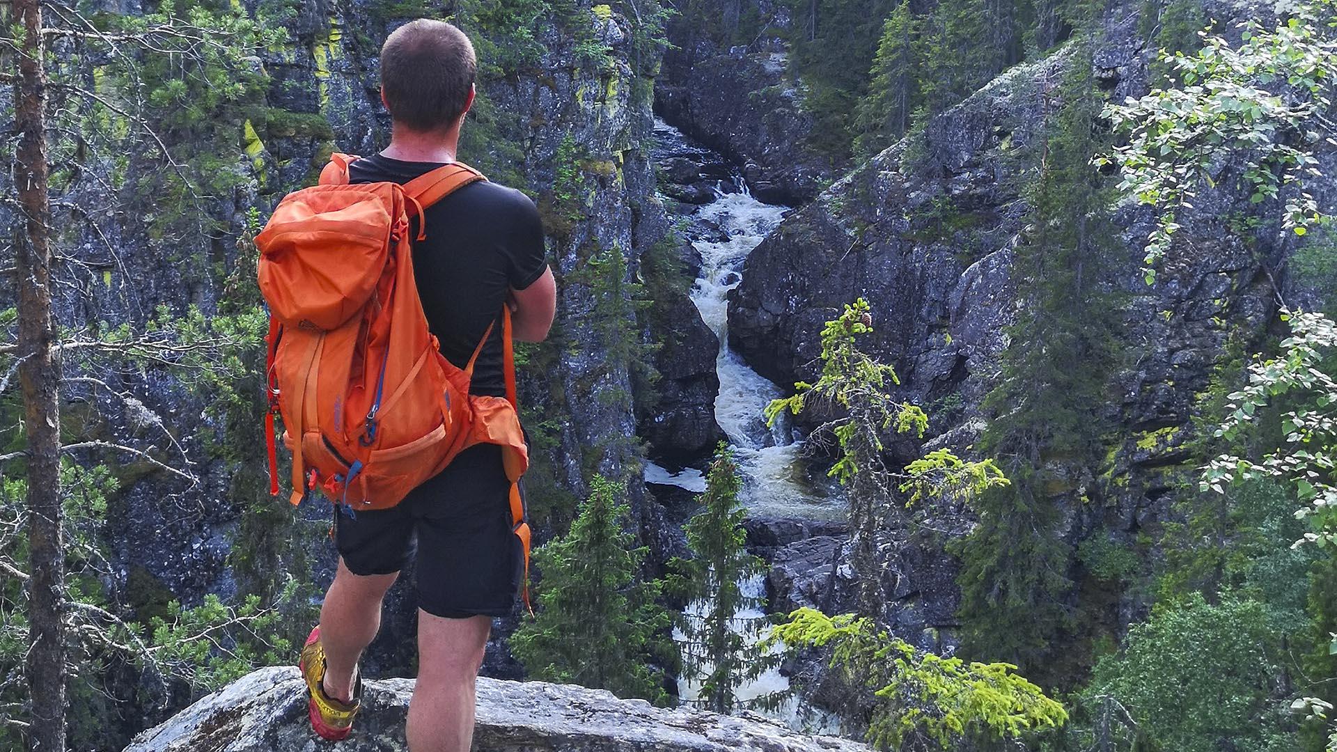 Ein Mann mit orangem Rucksack steht an einem Aussichtspunkt über einer Flusschlucht in einem Wald