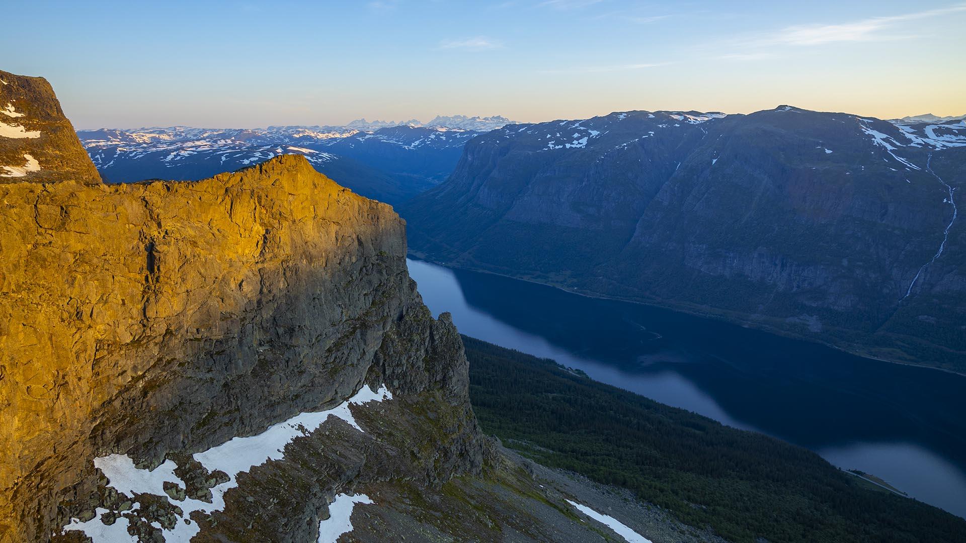 Utsikt mot bratte fjell med litt snø i fjellsidene og en innsjø deler fjellene.