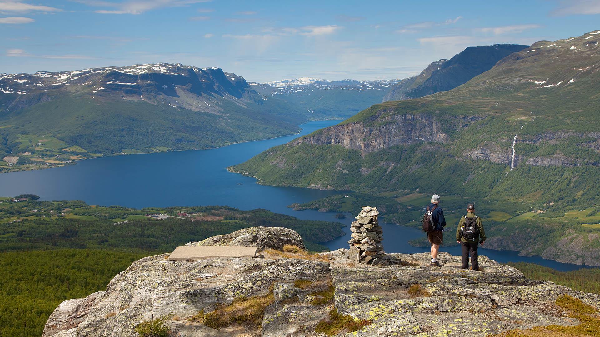 Two persons on top of a mountains standing beside a cairn. View towards large lake surrounded by mountains.