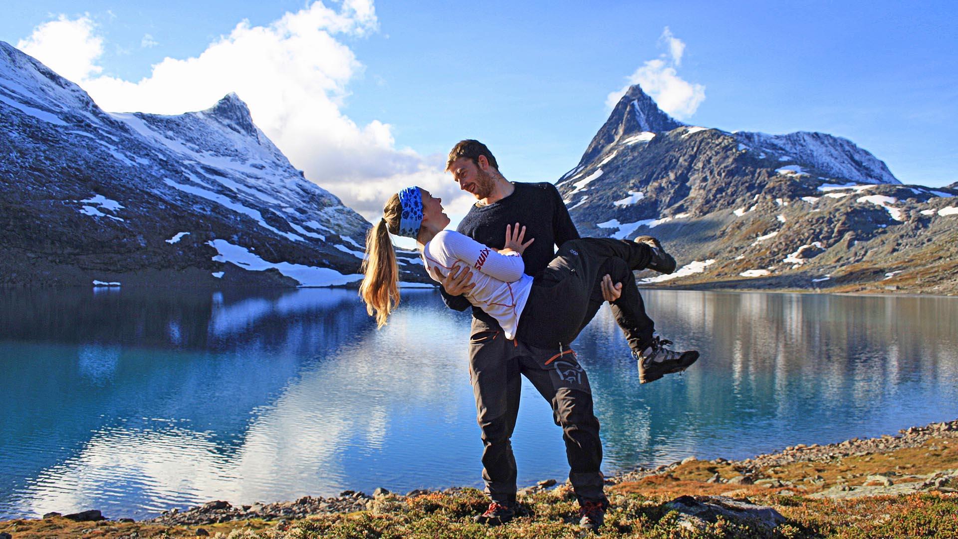 Mann holder kvinne i armene foran en innsjø med spisse tinder i bakgrunnen.
