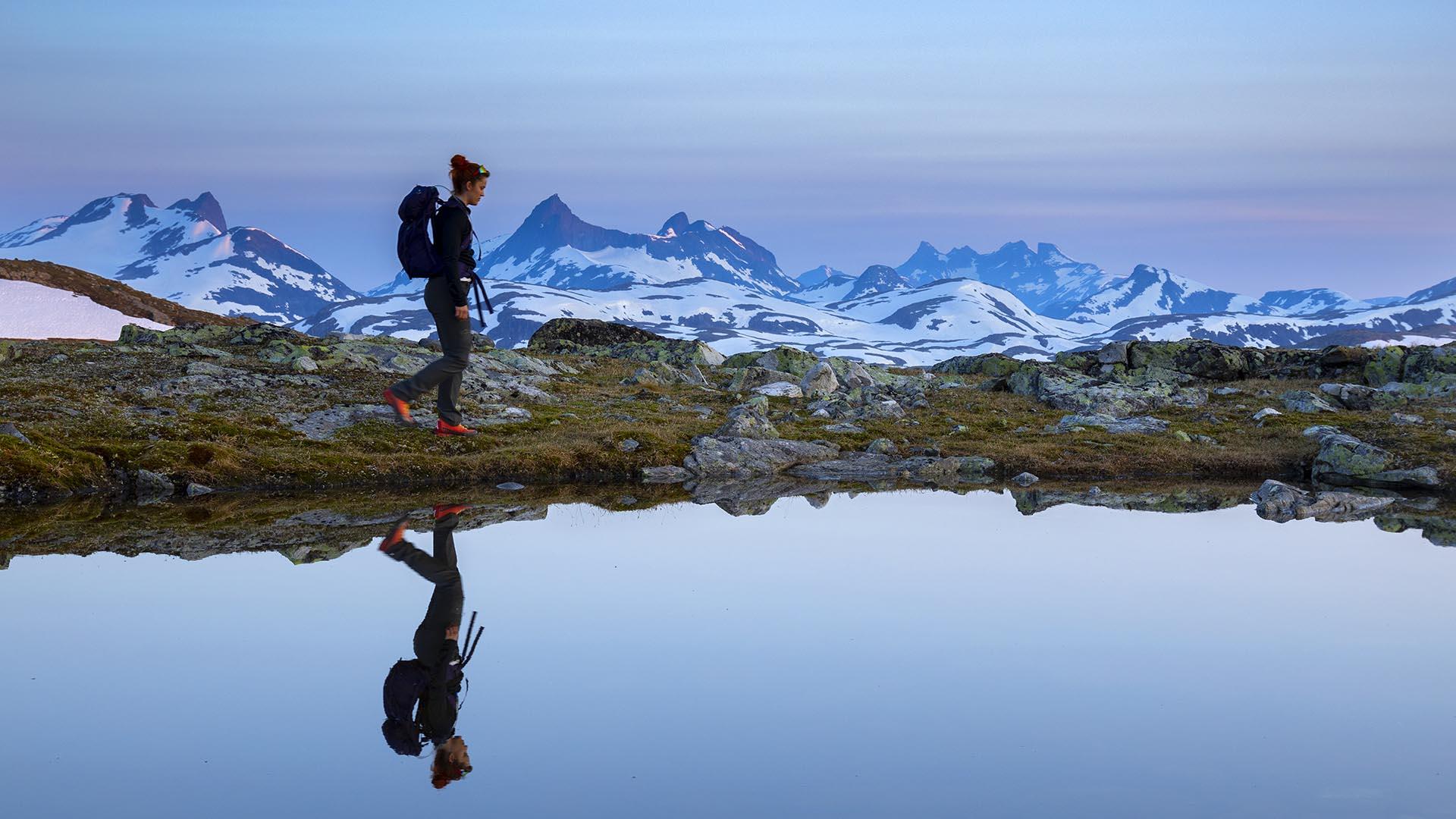 Crystal clear lake, woman walking by the lake is reflected by the water surface. High, pointed mountains in the far background. Purple morning light before sunrise.