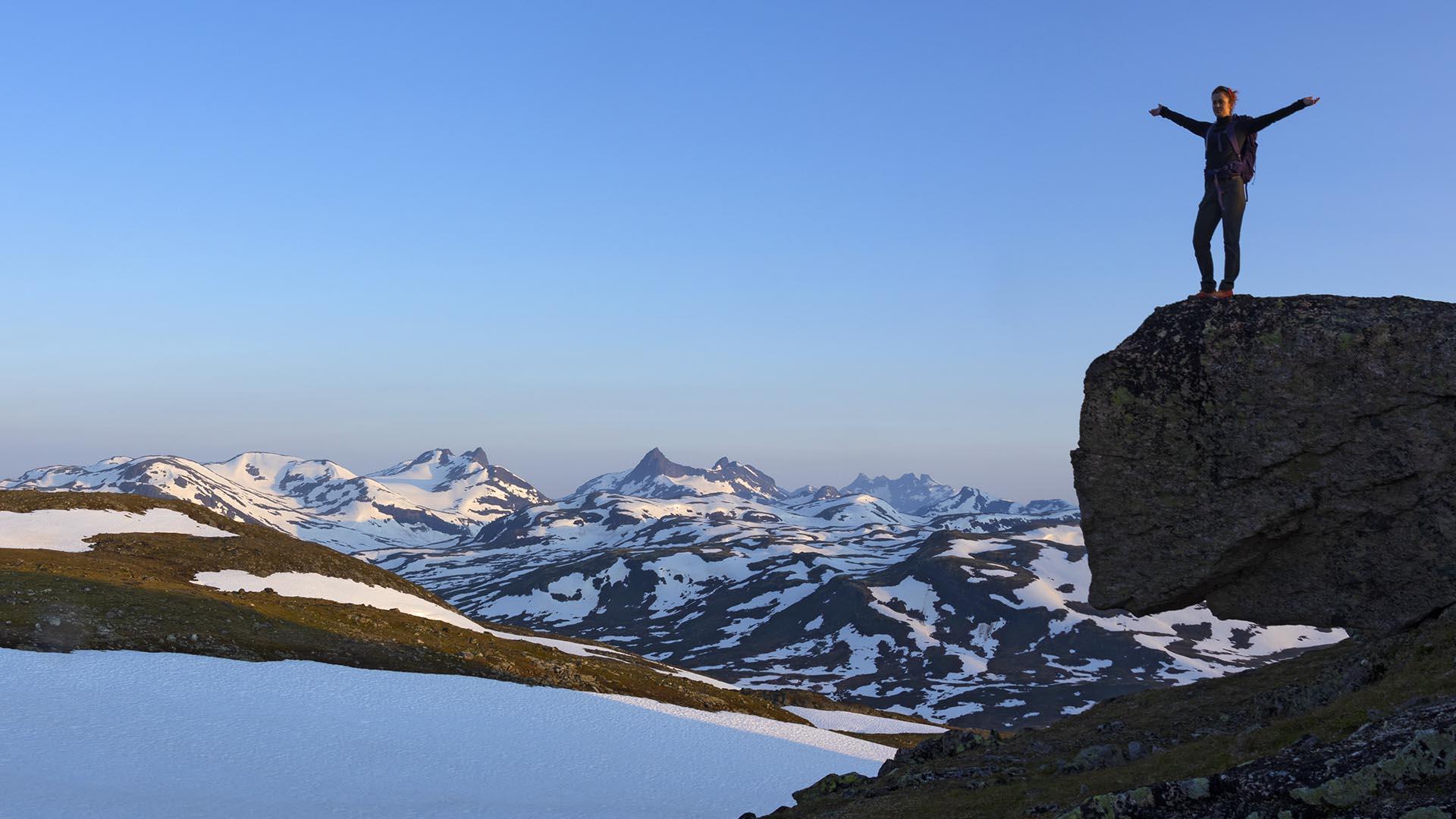 Vandrer på toppen av stein til høyre bildet nyter utsikt over snøflekkede ville fjell i bakgrunnen en skyfri dag.