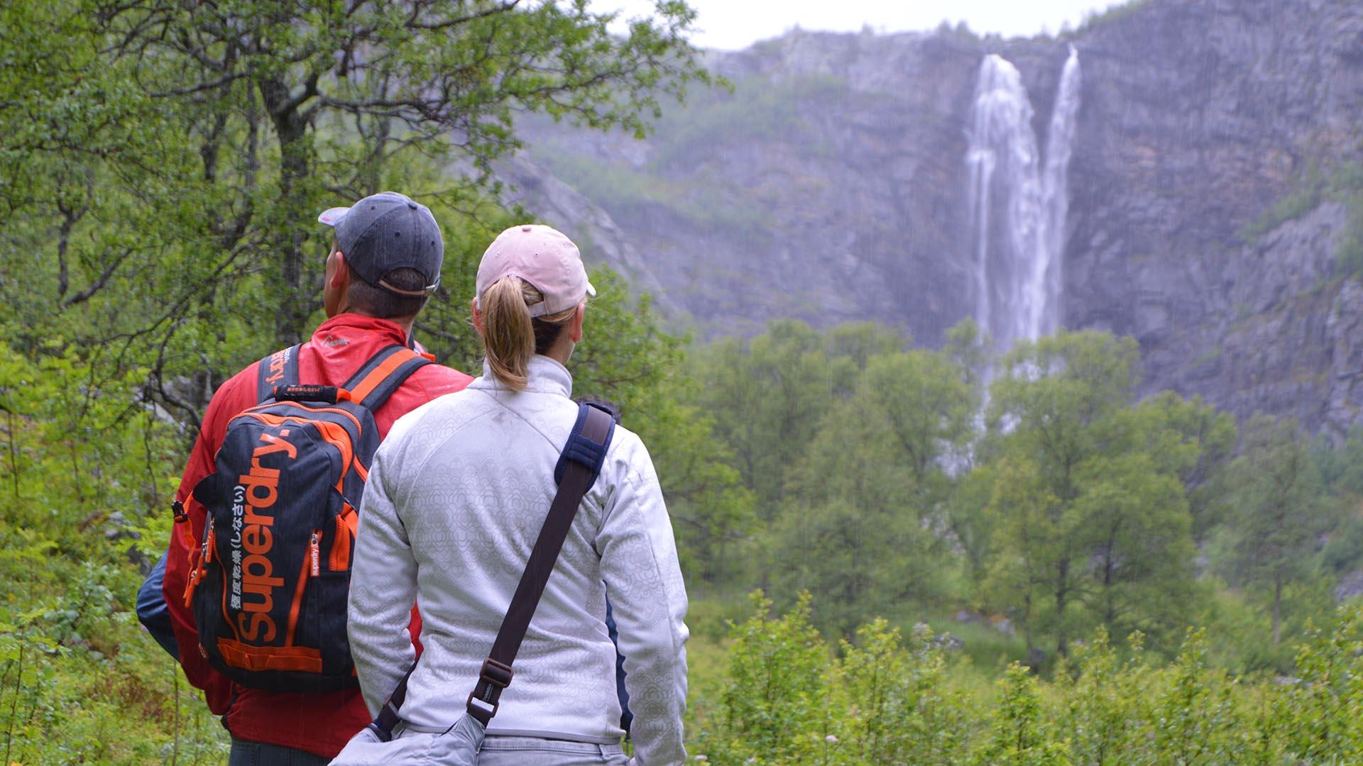 Two hikers are standing in a lush valley. In the background there is a rock cliff from which a waterfall plunges.