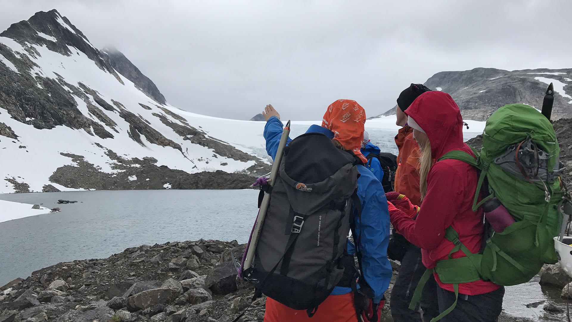 Mountain hikers discuss the route. One points towards a glacier and a high, pointed peak betond a lake.