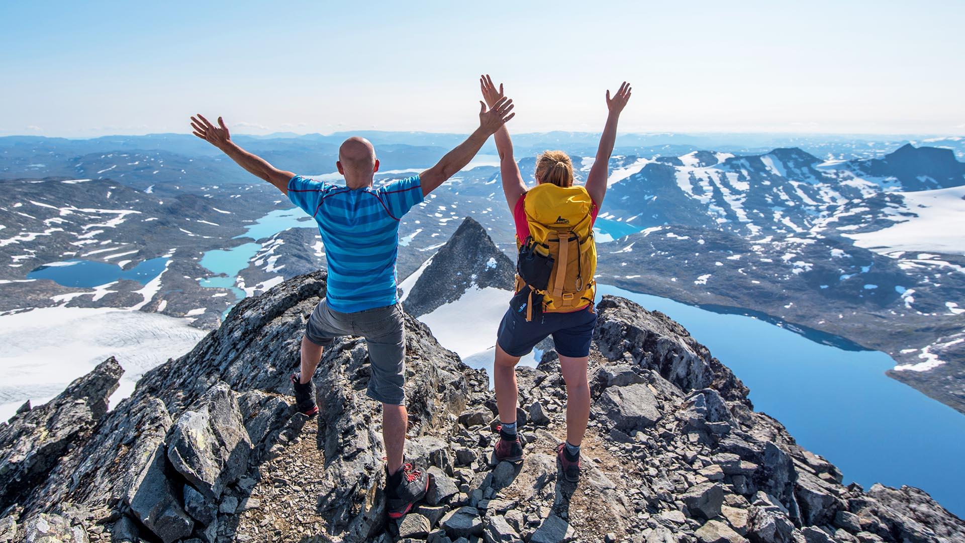 To fjellvandrere i sommerklær på toppen av et høyt fjell med armene i været idet det skuer over et goldt høyfjellslandskap med breer, vann og andre høye fjell.