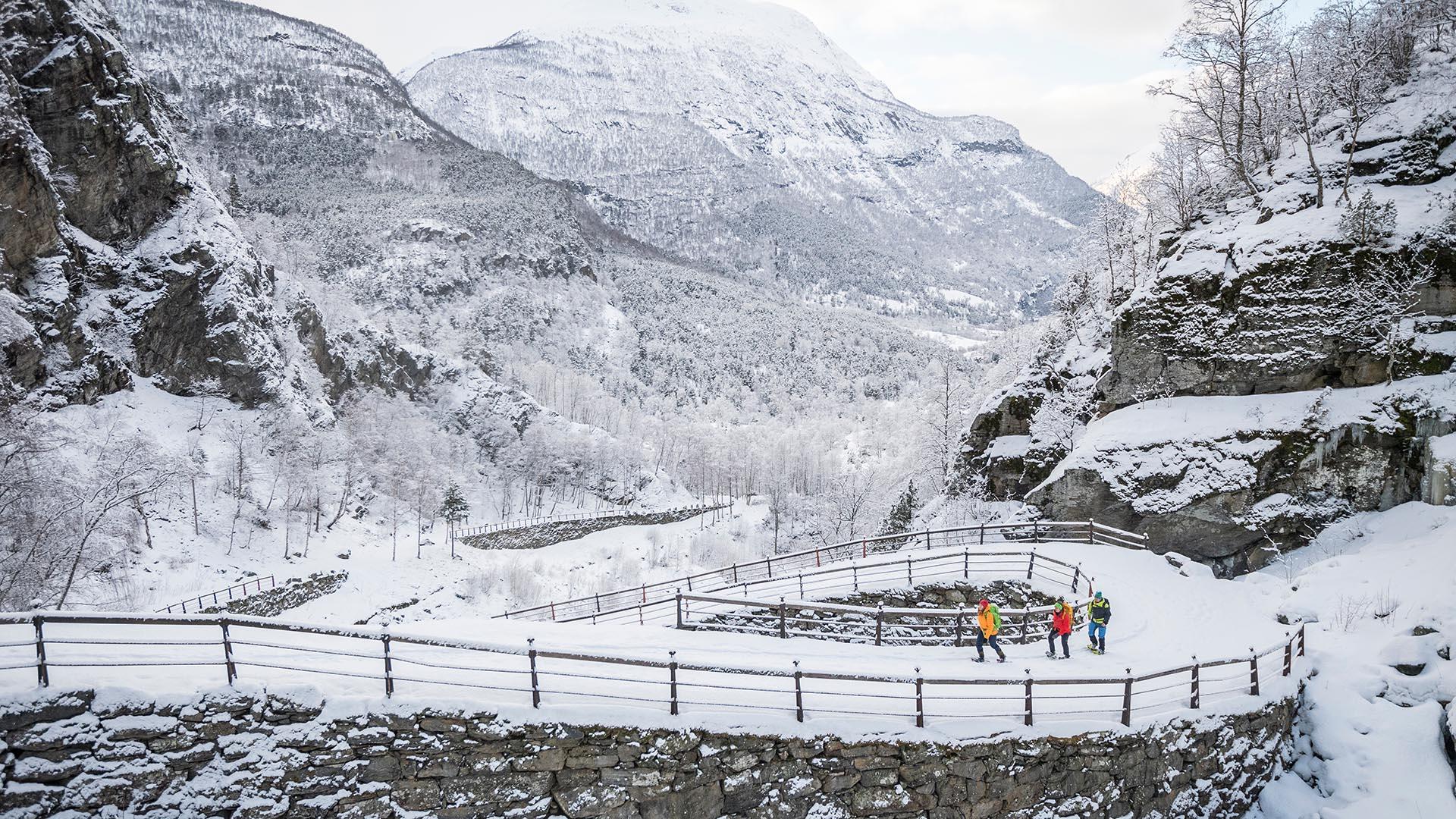 Three persons in colourful clothes on snowshoes walk a historical road that winds up a hill, surrounded by steep mountain slopes in a black and white winter landscape.