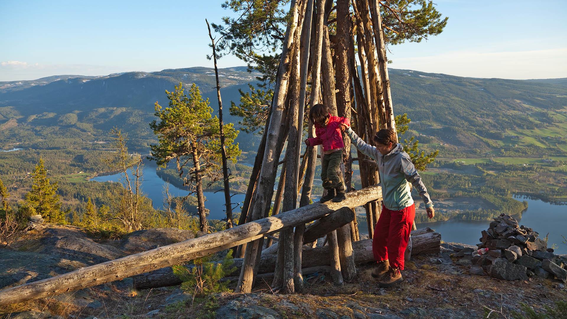 Barn som balansere på tømmerstokk som har falt ned fra en varde på en fjelltopp. Utsikt mot innsjøer og fjell i bakgrunnen