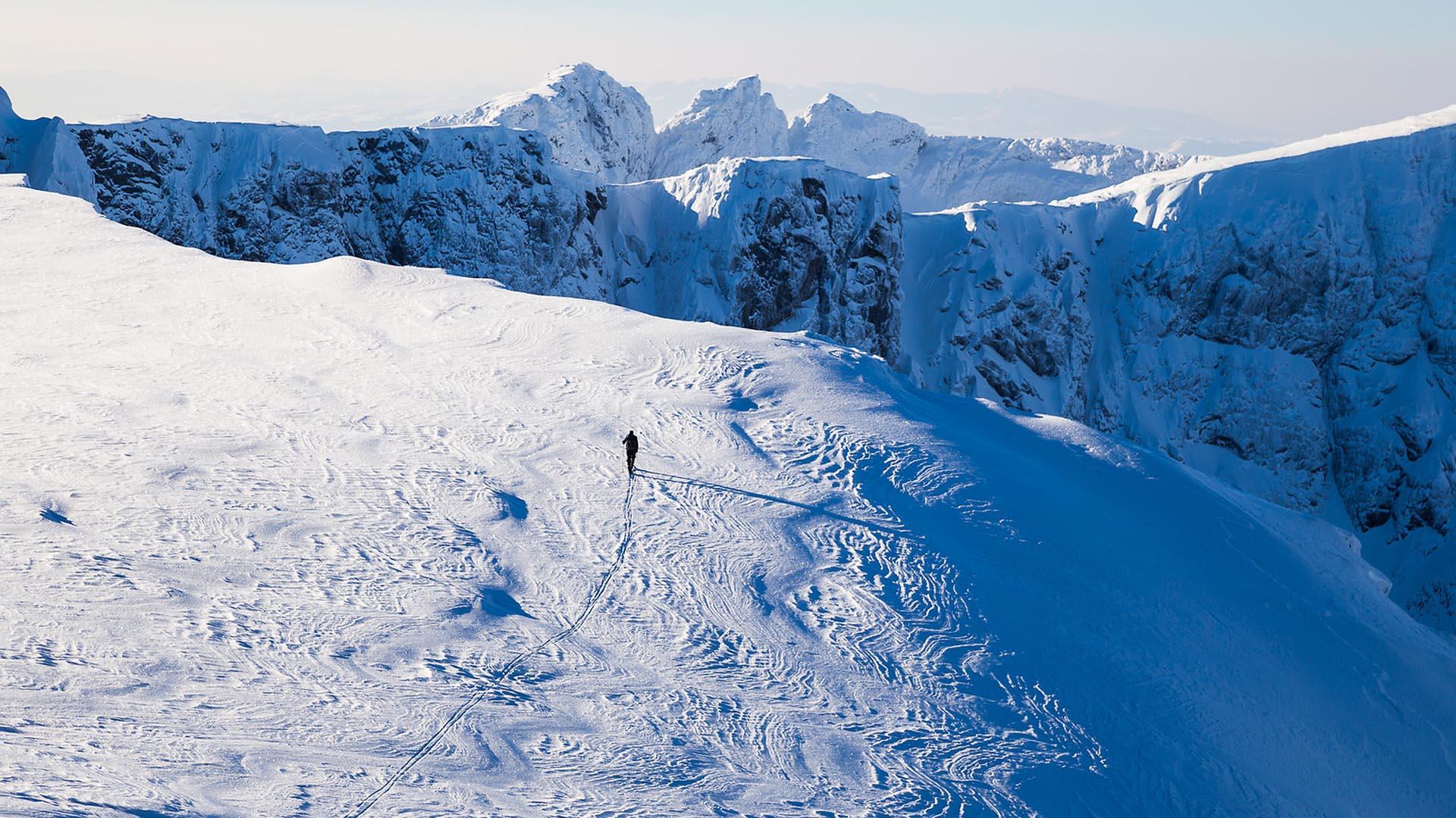 person på fjellskitur godt over tregrensen sett fra et stykke unna. Ville fjelltopper i bakgrunnen.