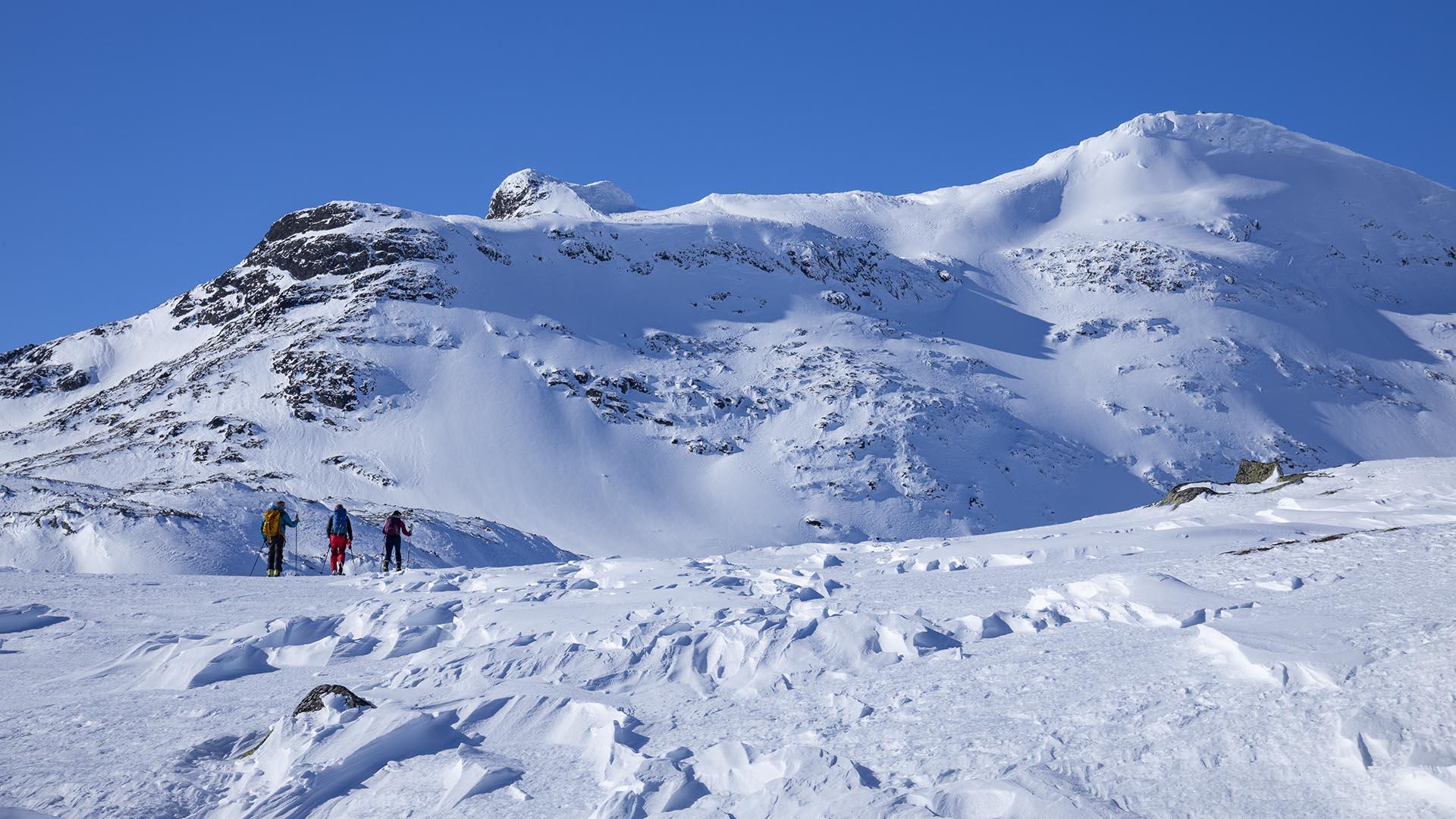 From a distance we see three people on the mountains skiing towards a mountain top we see in the background.