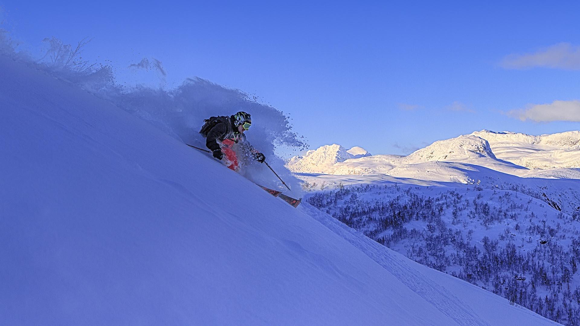 Person on its way down a hillside on skis with mountains in the background.