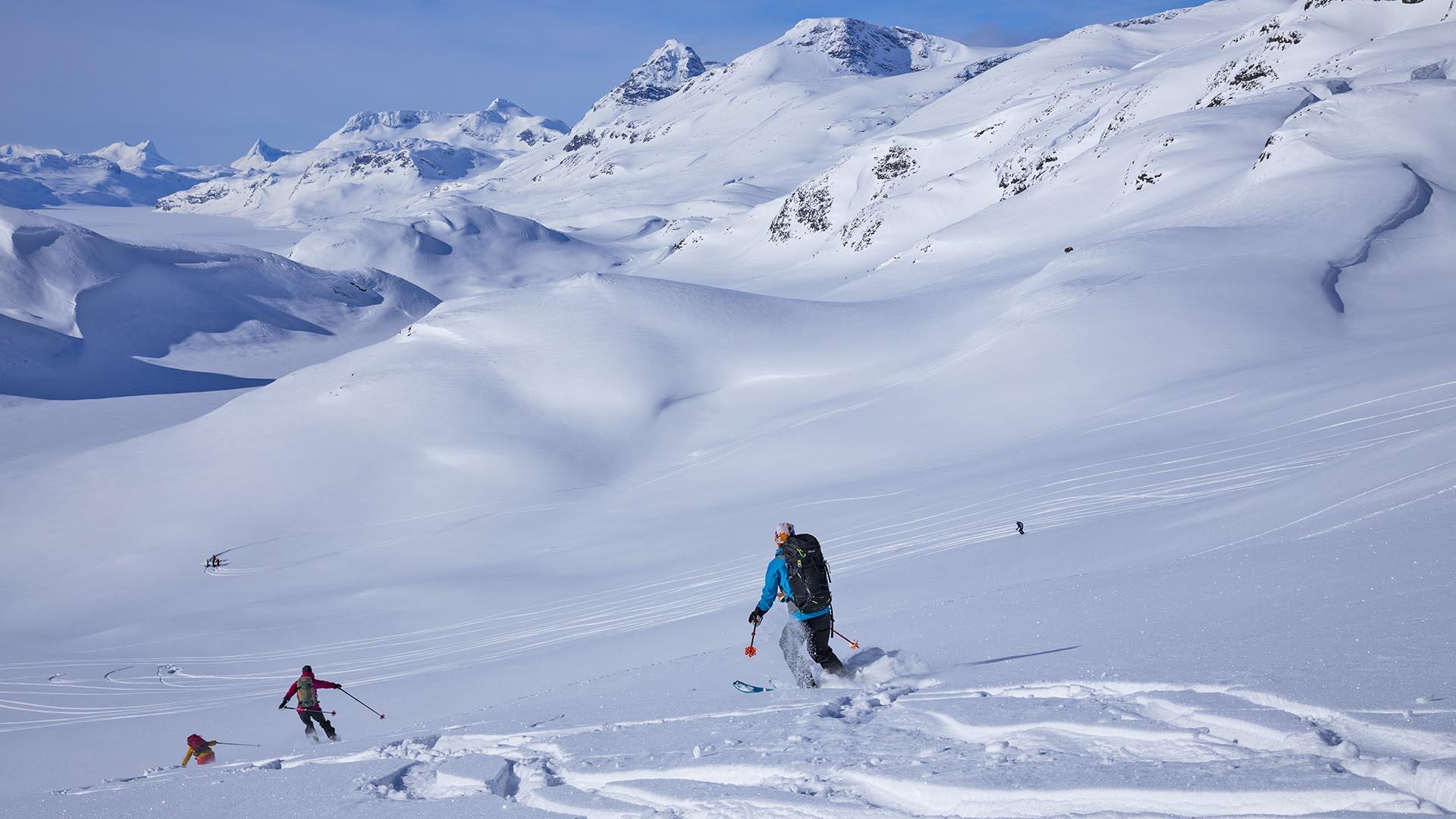 3 personer på vei ned fjellside med randoneeski. Snødekte spisse fjell i bakgrunnen.