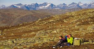 Eine Familie macht eine Pause im Gras zwischen flechtenbewachsenen Steinen auf einer Hochebene an einem sonnigen Herbsttag. Am Horizont hohe, spitze Berge.