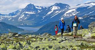 Hikers in the mountain pass a section with a lot of lichen-covered rocks. A lake and high mountains in the background.