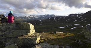 En person sitter på en stor stein og ser ut over fjellandskap i motlys
