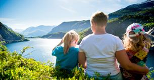 En familie sitter i en li og nyter utsikt over vann og fjell.