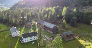 Dronenfoto des Hofes mit Gebäuden, großen Rasenflächen und angrenzendem Birkenwald. Sonnenstrahlen im gegenlicht erzeugen ein Mosaik aus Licht und Schatten.
