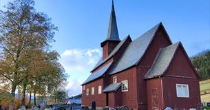 A red-brown stave church surrounded by a church yard with birch trees that start to get autumn-coloured leaves.