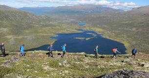 Fjellvandrere i rad og rekke med vann som ligger i dalen i bakgrunnen.