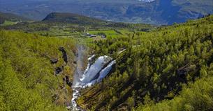 En dobbel foss som spruter mye vann. Bygd og fjell i bakgrunnen.