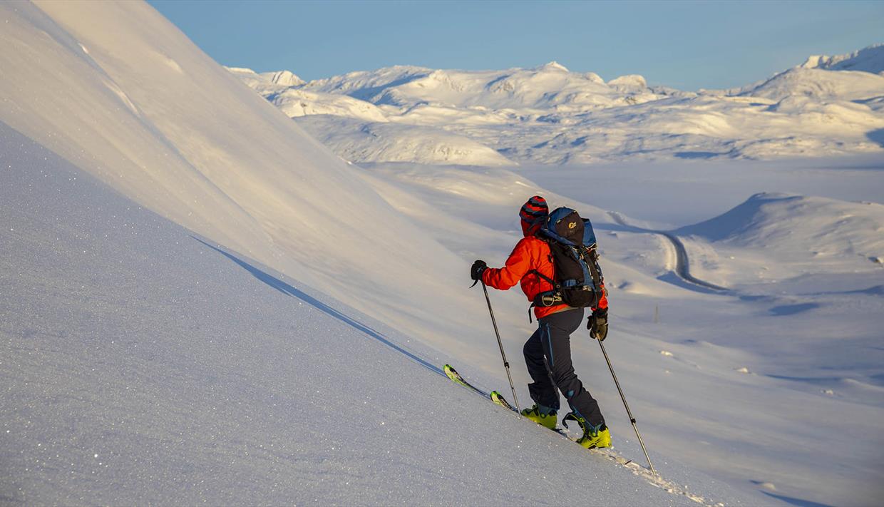 Person på vei opp fjellside med randoneeski, vid utsikt over snødekt fjellandskap