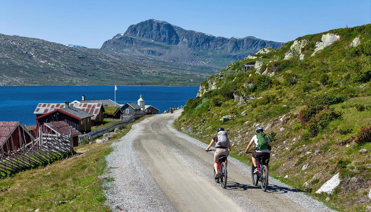 To syklister på grusvei langs vann med fjell i bakgrunnen.