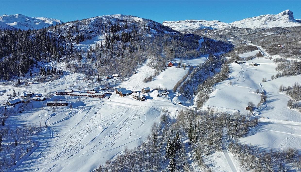 Luftfoto einer Mountain Lodge in einem sonnigen, schneebedeckten Hang, umgeben von Wald und hohen Bergen