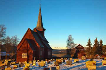 En stavkirke i en snødekt kirkegård i vintersolskinn.