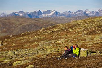 En familie tar en pause i gresset på et fjellplatå mellom mange lavdekte steiner på en solrik høstdag. Høye, spisse fjell i bakgrunnen.