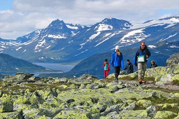 Hikers in the mountain pass a section with a lot of lichen-covered rocks. A lake and high mountains in the background.