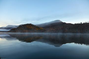 Der Nøsakampen und sein Spiegelbild vor Sonnenaufgang vom Kajak auf dem Helin aus gesehen. Frostnebel hängt über der stillen Wasseroberfläche, und das Fjell leuchtet in Herbstfarben.