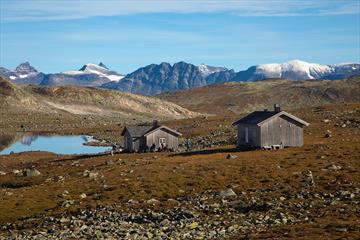 Tomashelleren en flott høstdag. To gråfargede hytter ved et lite vann, og ruvende fjelltopper i bakgrunnen.