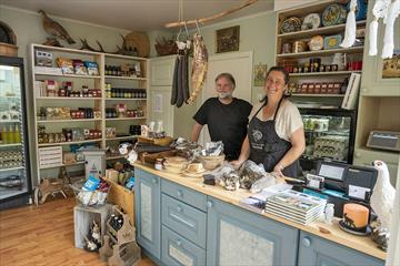 The owners of the store standing behind the counter of the small, cosy shop.The shelves are filled with delicious local food.