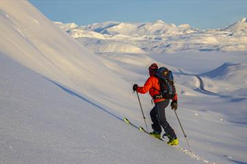 Person på vei opp fjellside med randoneeski, vid utsikt over snødekt fjellandskap