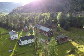 Dronebildet av gården ovenfra med flere bygg, store plenarealer og tilgrensende skogsholt, og solstråler som skaper lys og skygge.