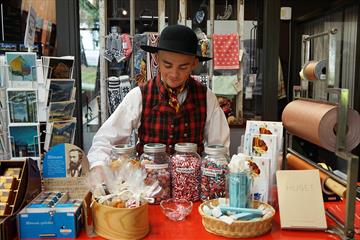 En bunadkledd ung mann bak disken på en museumsbutikk med gammeldagse drops på glass, strikkede votter, kunstkort og bøker.