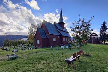 Stavkirke på grønn plen med et lite rognebærtre i motsol.