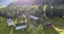 Dronebildet av gården ovenfra med flere bygg, store plenarealer og tilgrensende skogsholt, og solstråler som skaper lys og skygge.
