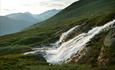 A waterfall in a green hillside with high summits in the background.