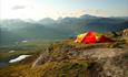 Ein gelb-rotes Zelt auf einem grasigen Absatz mit Panoramaaussicht zu hohen Bergen.