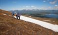 Hikers on a grassy mountain slope with snow fields. A lake and high mountains in the background